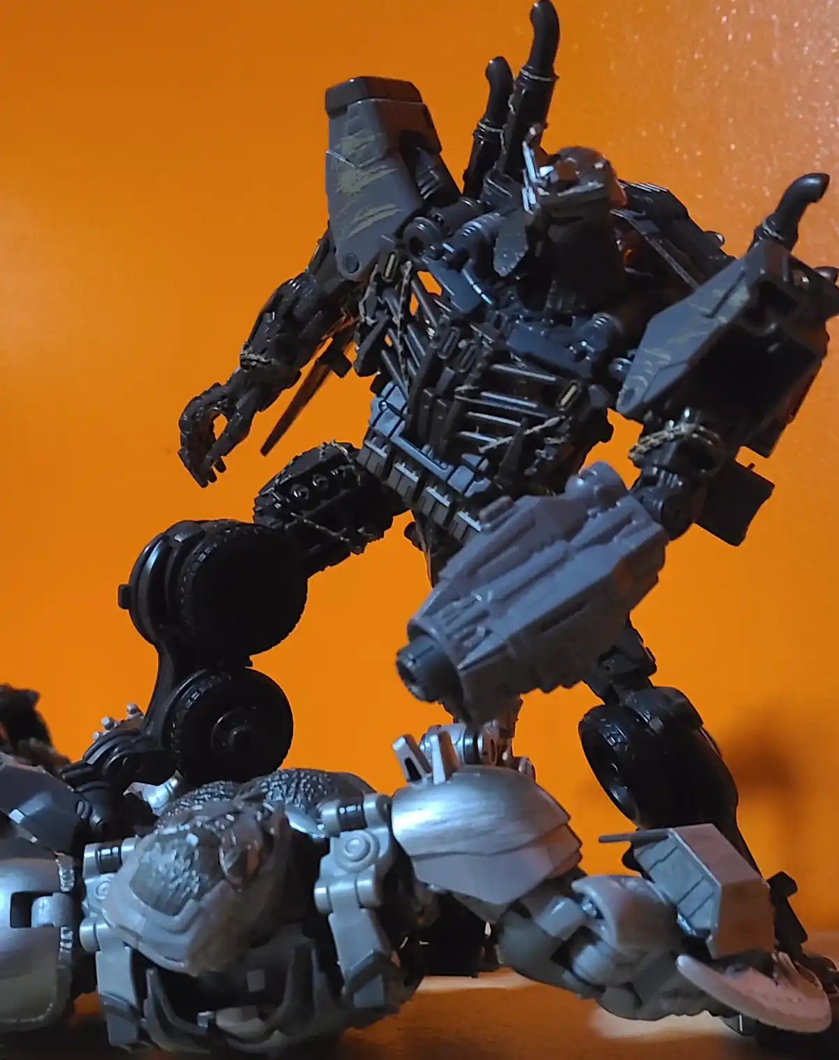 A black, heavily detailed transformer toy stands over a defeated silver transformer toy against an orange background. The…