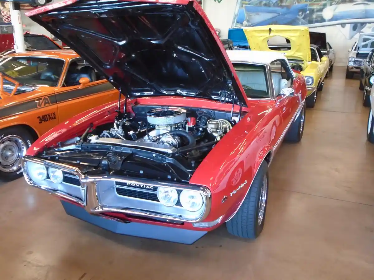 A red classic Pontiac Firebird with its hood open, revealing a detailed engine, is displayed in a car showroom alongside…