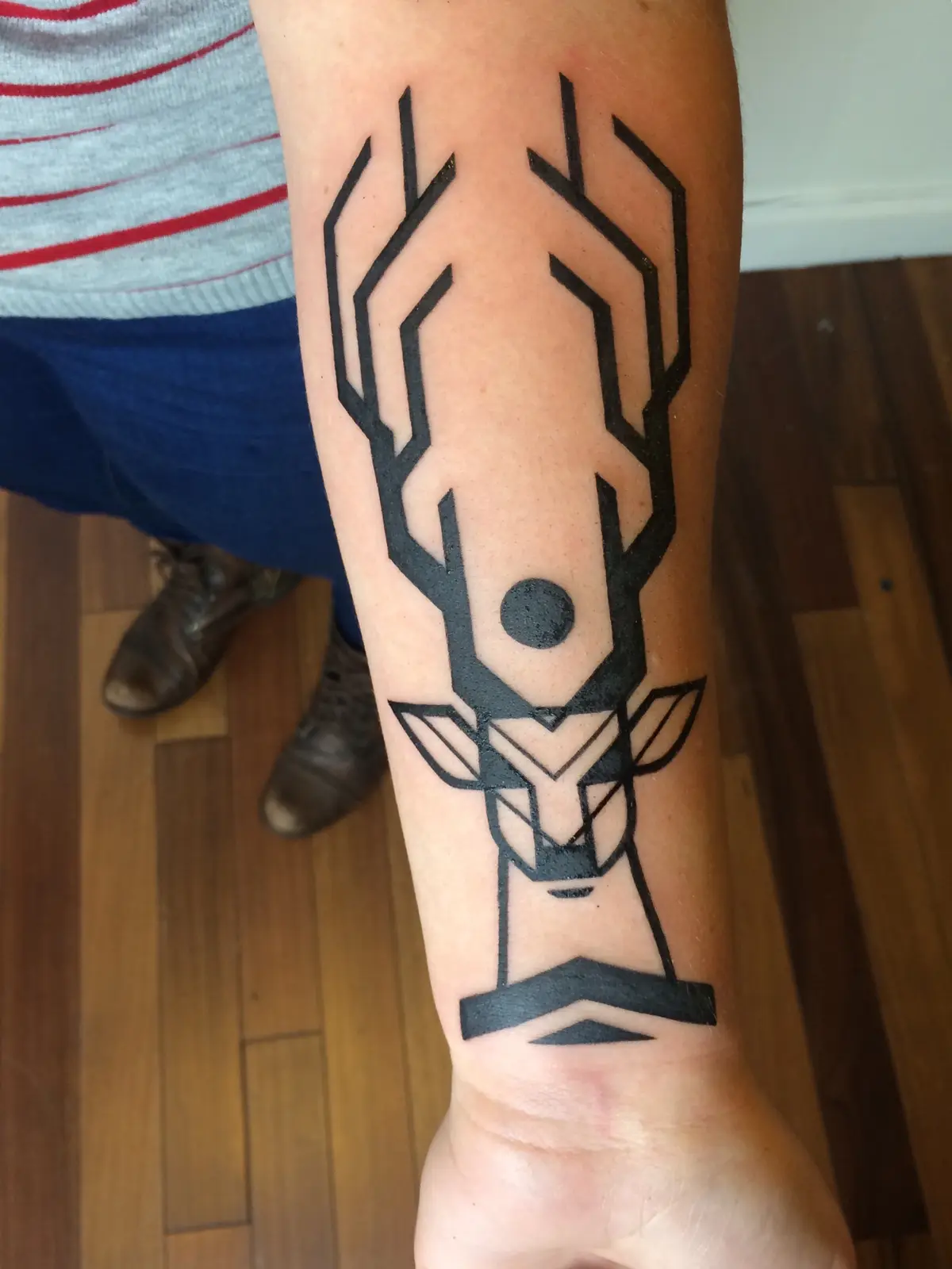 A close-up photo shows a person's forearm with a black geometric tattoo of a stylized deer head, featuring angular antlers…