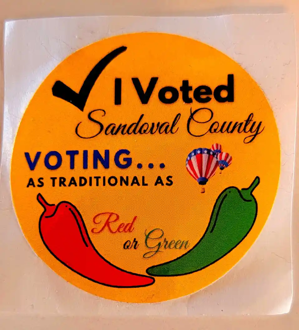 A circular yellow 'I Voted' sticker from Sandoval County features a black checkmark, two chili peppers (one red, one green)…