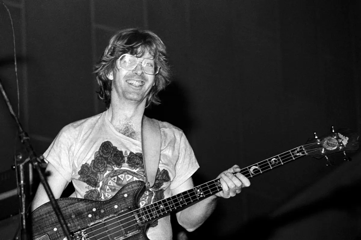 A black-and-white photo shows a smiling man with glasses and shoulder-length hair playing a bass guitar on stage. He wears a…