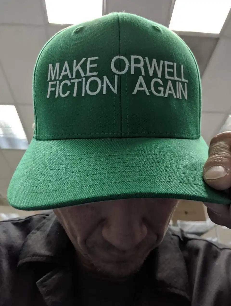 A person wearing a green baseball cap with white embroidered text that reads 'MAKE ORWELL FICTION AGAIN', holding the brim…
