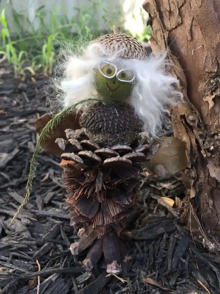 A whimsical handmade figurine crafted from natural materials sits beside a tree trunk. The figure has a green acorn cap for…