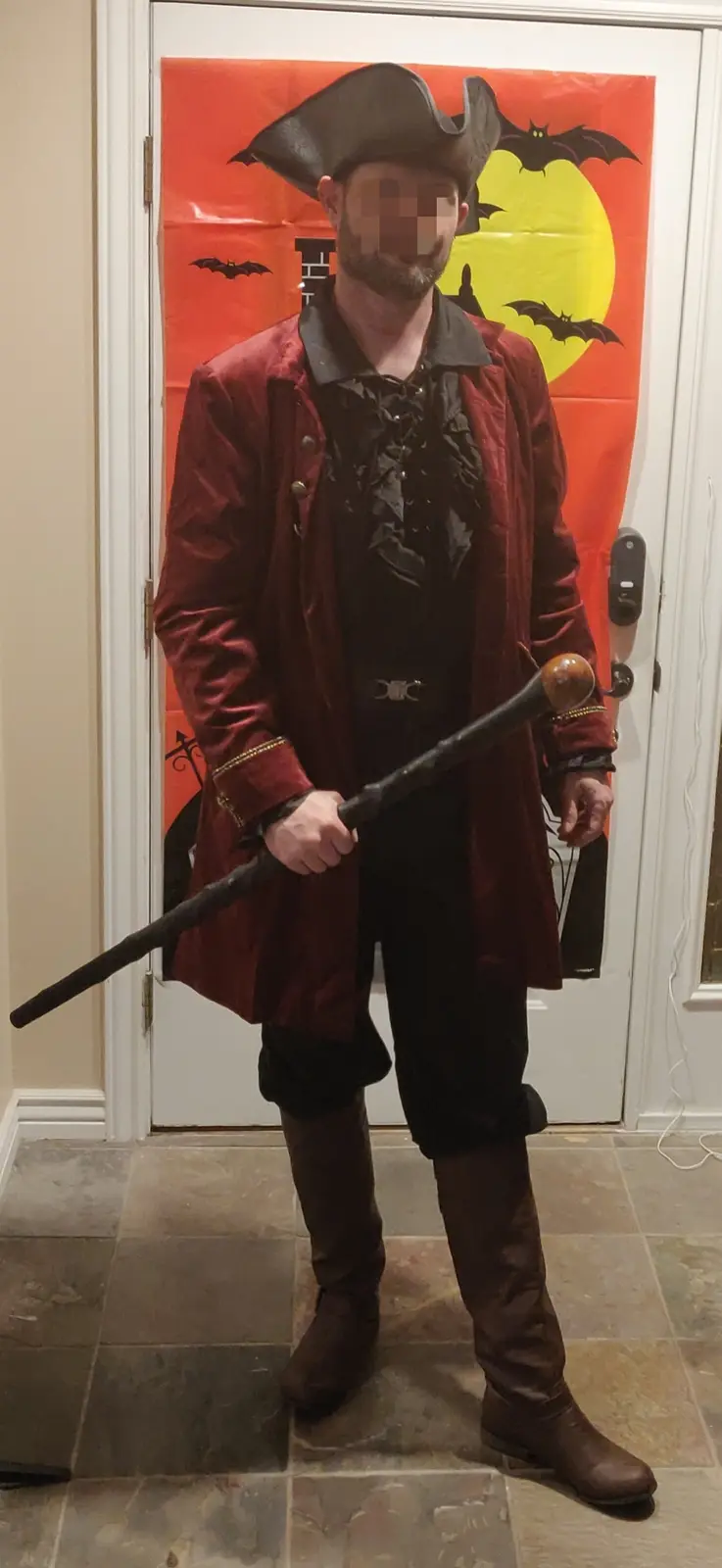A man in a pirate costume, featuring a tricorn hat, red coat, and brown boots, stands in front of a Halloween-themed door…