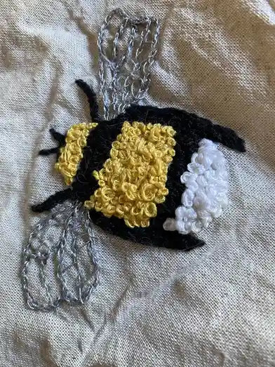 A handmade bee crafted from black, yellow, and white yarn with silver wire wings, stitched onto a beige fabric background…