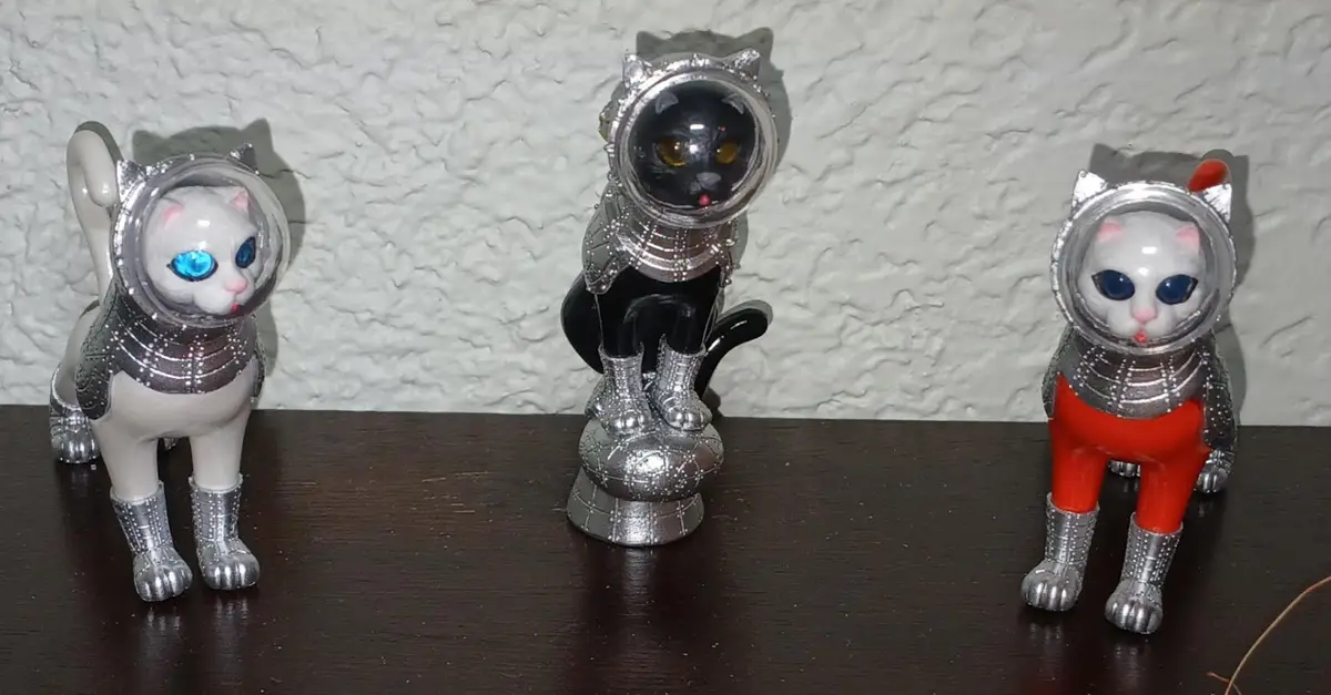 Three plastic cat figurines dressed in detailed silver astronaut suits with clear bubble helmets, standing on a dark wooden…