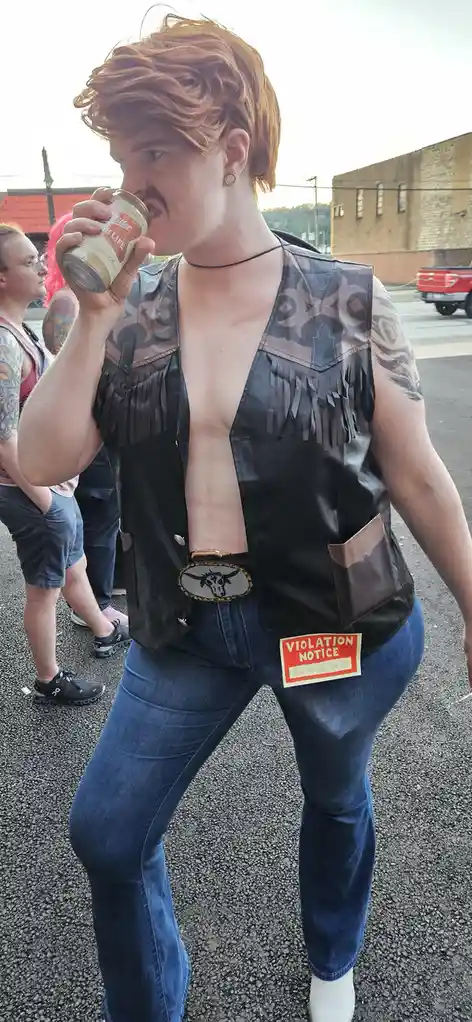 A person with red hair wearing a black fringed leather vest and jeans is drinking from a Miller High Life can. A red and…