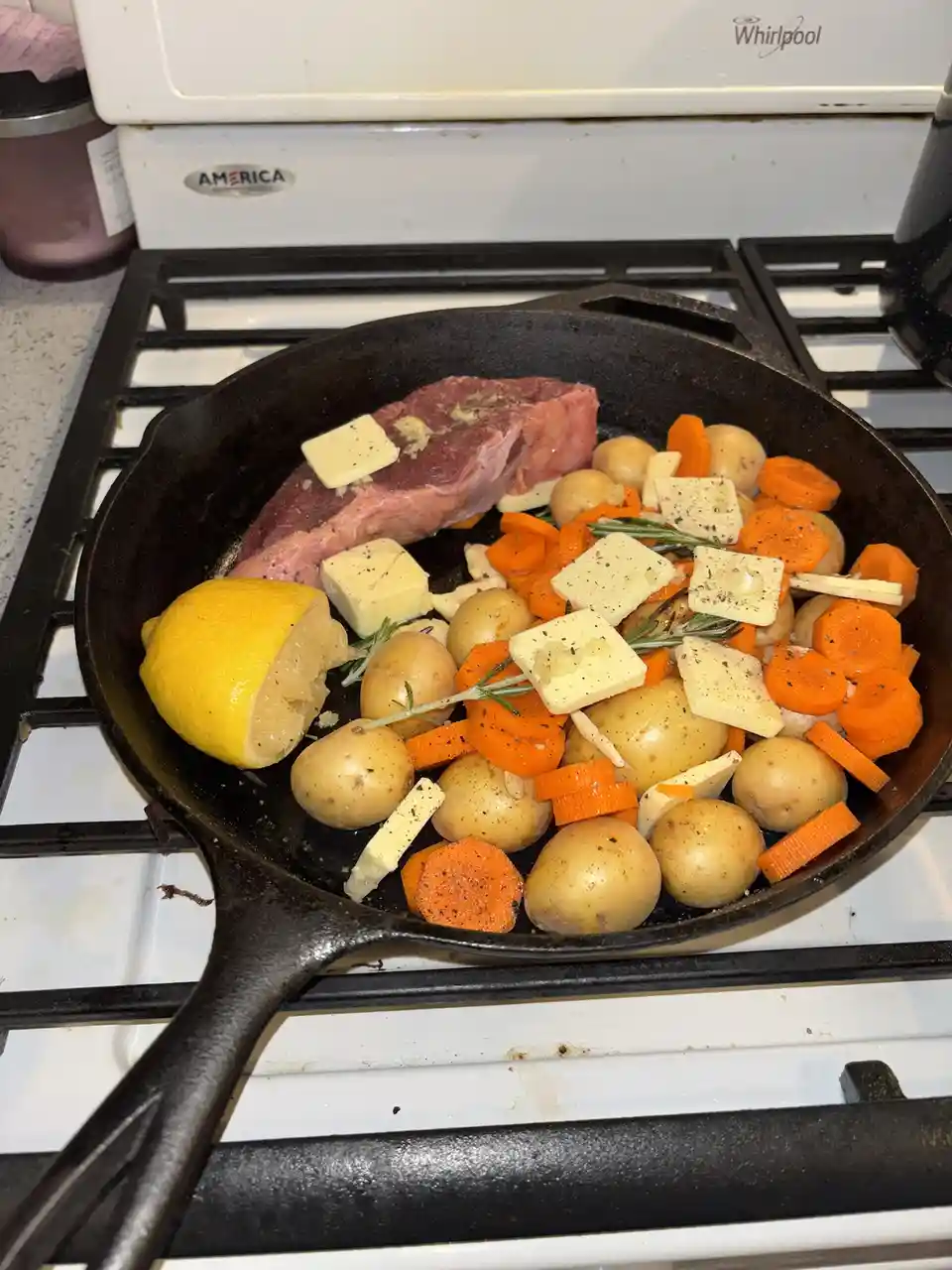 A cast iron skillet sits on a white Whirlpool stove burner, containing a raw steak, baby potatoes, sliced carrots, butter…
