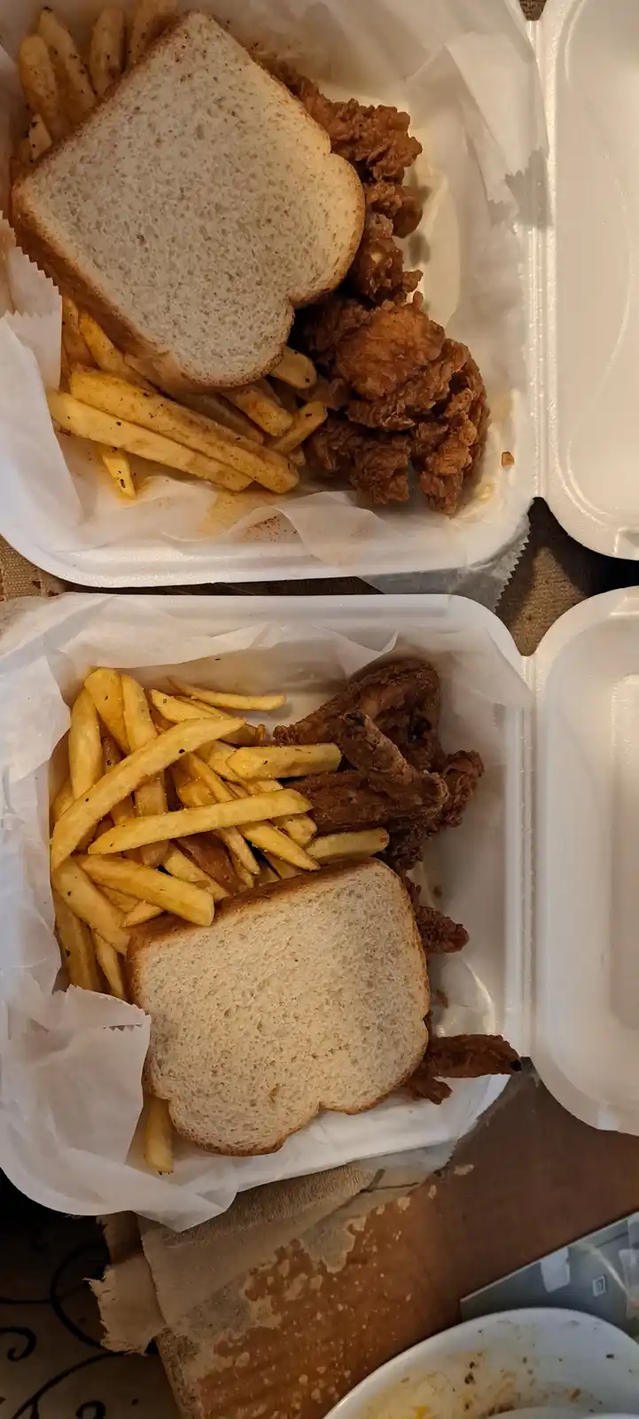 Two open white styrofoam takeout containers each hold a serving of fried chicken, french fries, and a single slice of white…