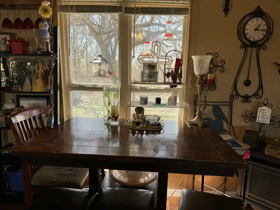 A cozy, cluttered dining nook with a wooden table, chairs, and various decorative items like candles, a clock, and bird…