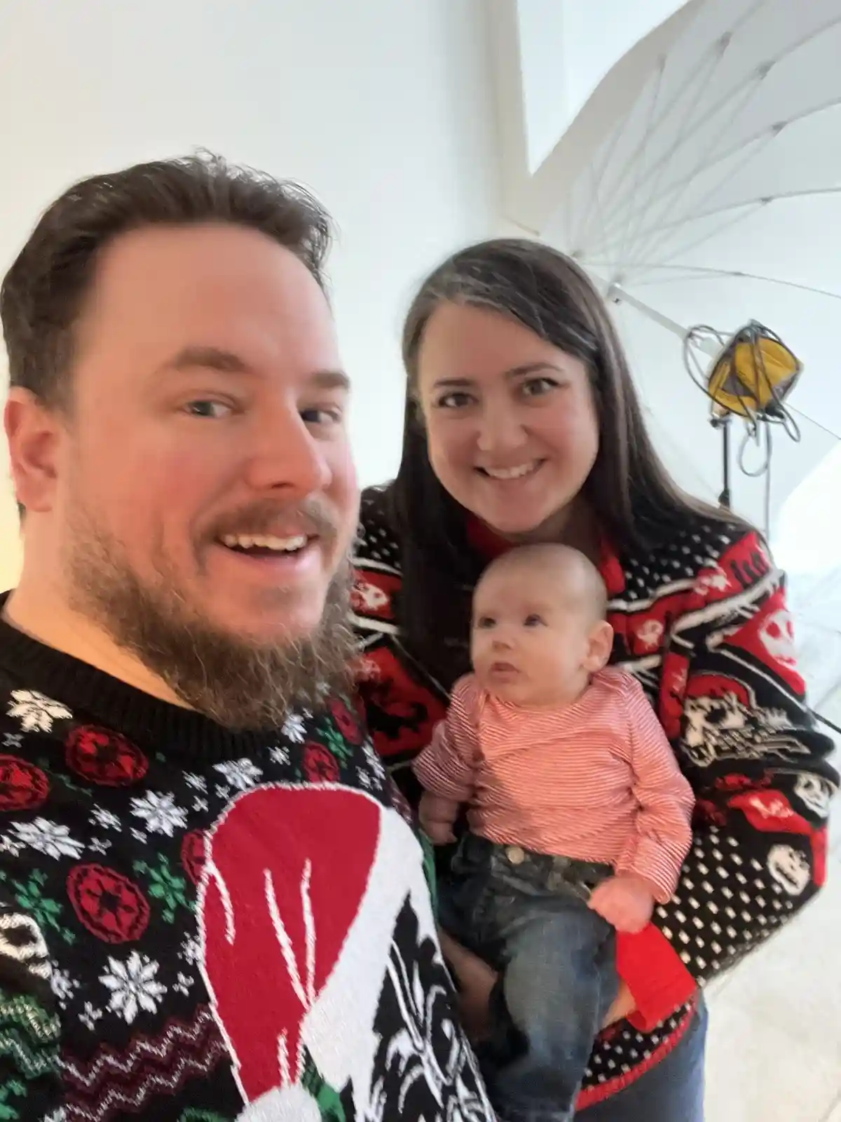 A man with a beard wearing a festive, overly decorated Christmas sweater smiles at the camera while holding a baby. Behind…