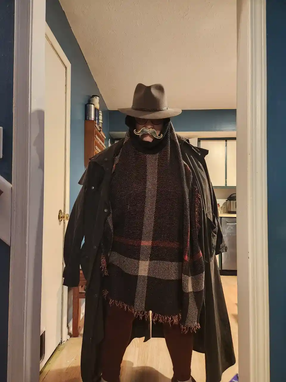 A person stands in a doorway wearing a wide-brimmed hat, glasses, a heavy black coat, and a large plaid scarf wrapped around…