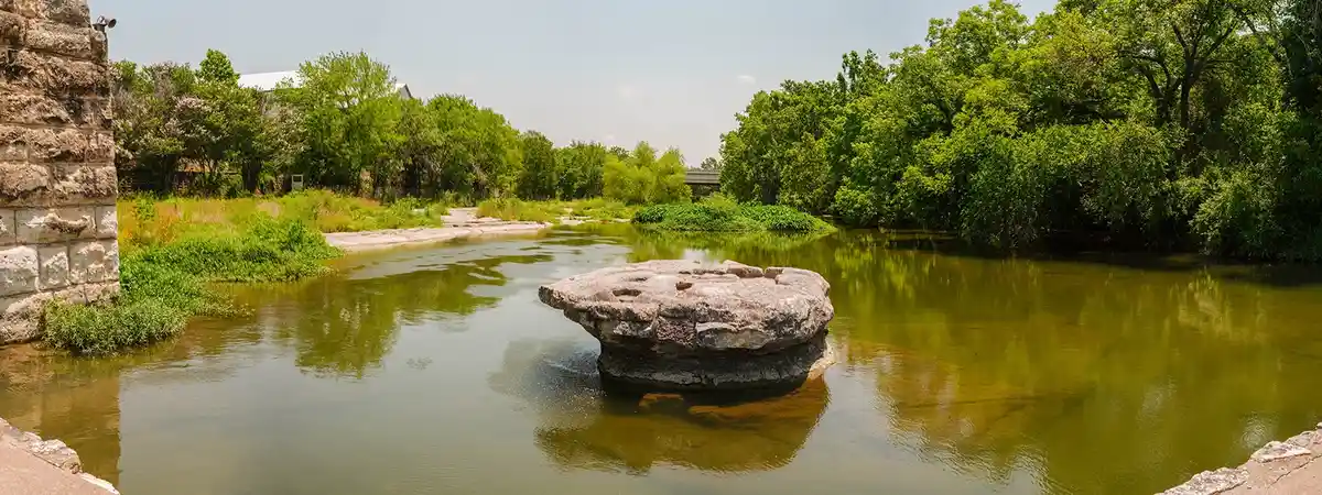 A wide-angle photo shows a calm, greenish river with a large, flat, circular rock in the middle. Lush green trees line the…