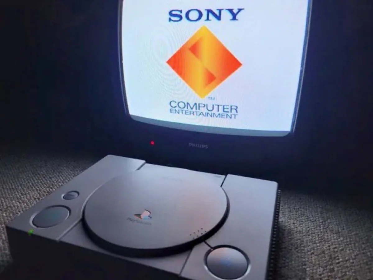 A silver PlayStation 1 console sits in front of a Philips CRT monitor displaying the Sony Computer Entertainment startup…