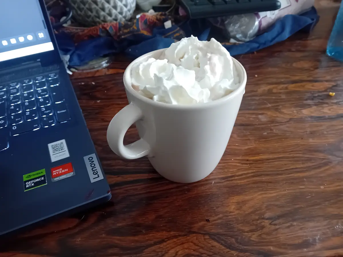 A white mug overflowing with whipped cream sits on a wooden desk next to a dark blue Lenovo laptop with visible NVIDIA…