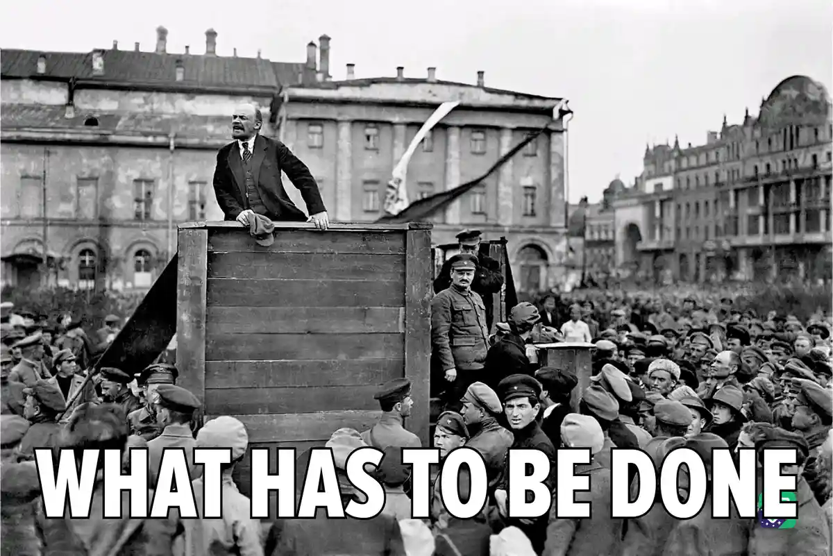 A black-and-white historical photograph shows Vladimir Lenin standing atop a wooden platform, addressing a large crowd of…
