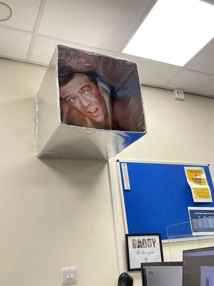 A silver duct tape cube mounted on a beige office wall features a photo of a man with a shocked expression peering out from…