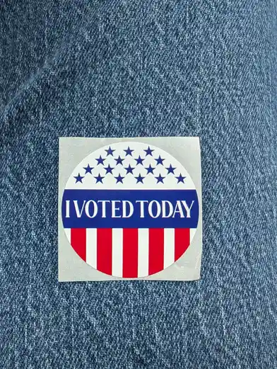 A circular 'I VOTED TODAY' sticker with stars and stripes design is stuck on a blue denim fabric. The sticker features a…