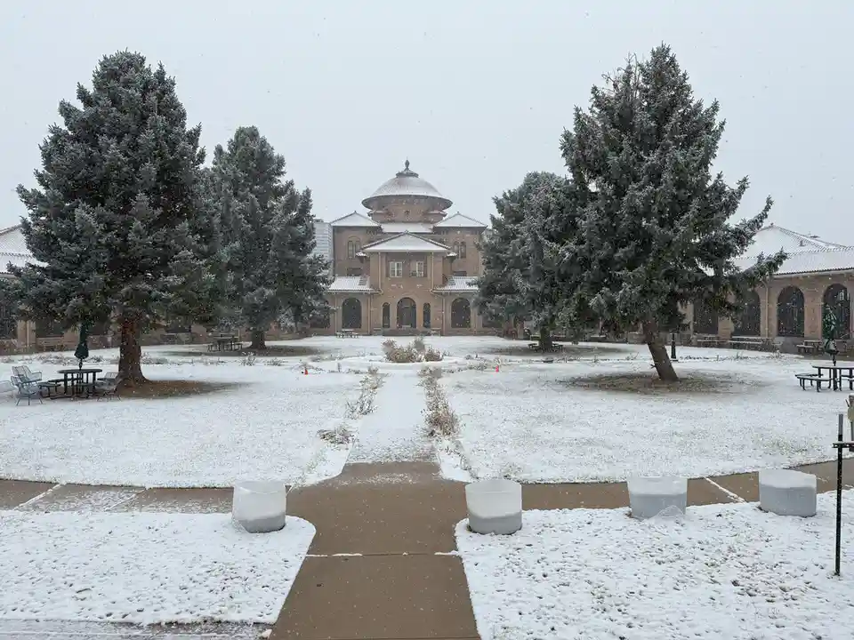 A snowy campus courtyard with a large domed building in the background, flanked by evergreen trees. The ground and pathways…