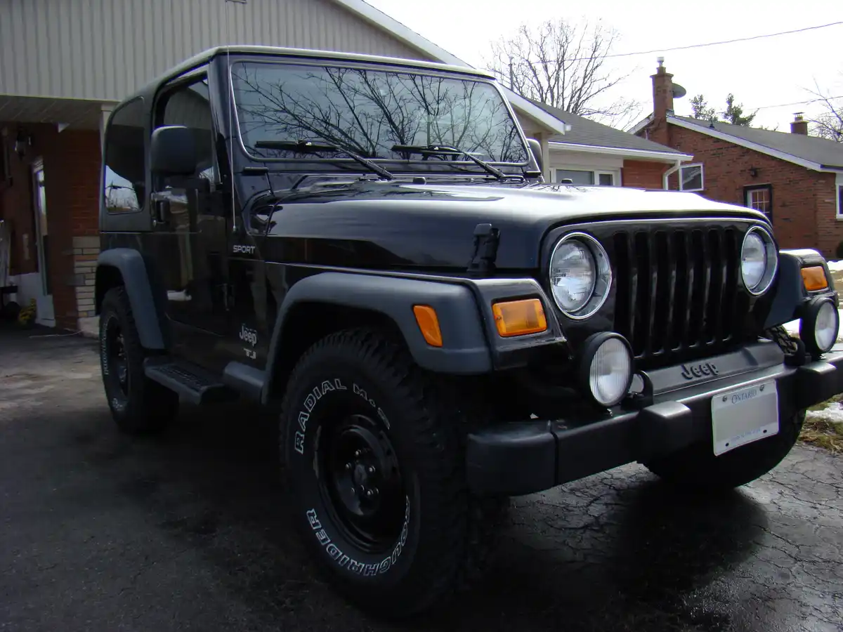 A black Jeep Wrangler TJ Sport is parked on a cracked asphalt driveway in front of a brick house with a metal-sided garage…