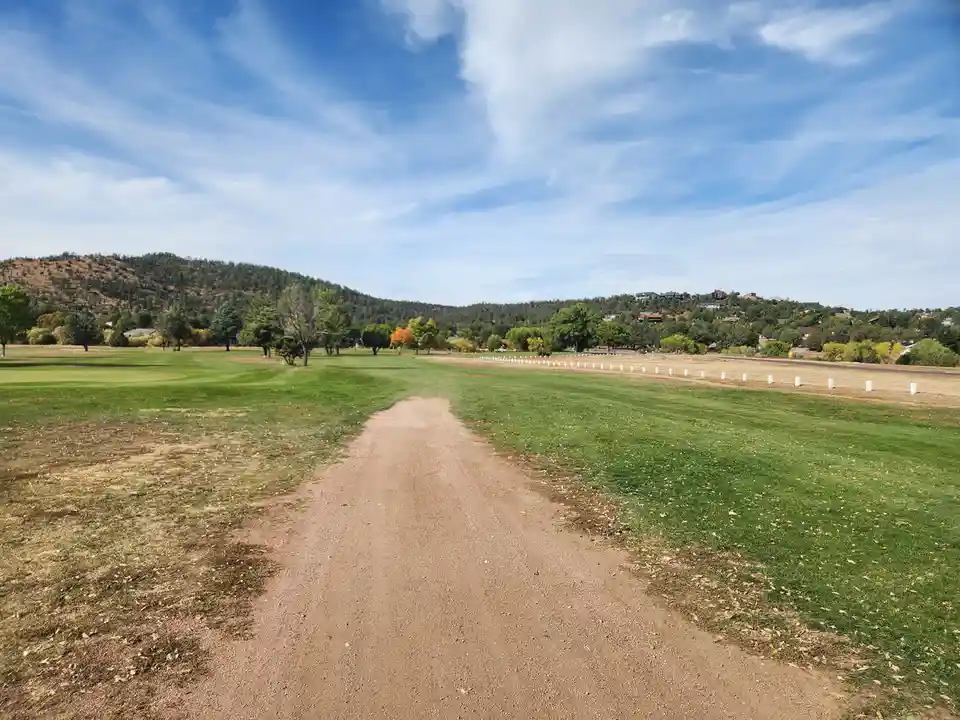 A wide dirt path cuts through a lush green golf course under a bright blue sky with scattered clouds. Trees and distant…