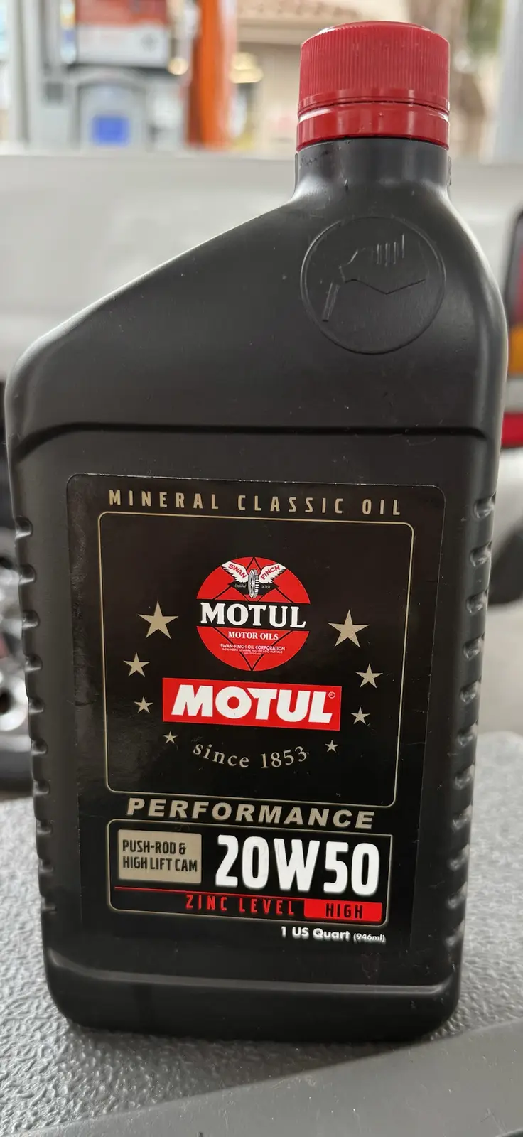 A black bottle of Motul mineral classic motor oil with a red cap sits on a gray surface. The label clearly displays 'MOTUL…