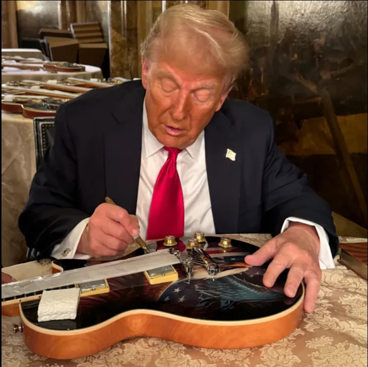 Former president Donald Trump is shown seated at a table, signing a guitar with a pen. He wears a dark suit, white shirt…