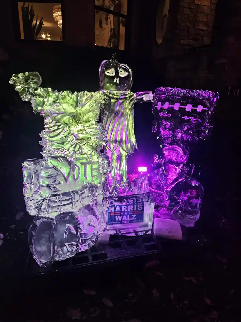An illuminated ice sculpture at night depicts three Halloween-themed figures: a green werewolf holding a 'VOTE' sign, a…