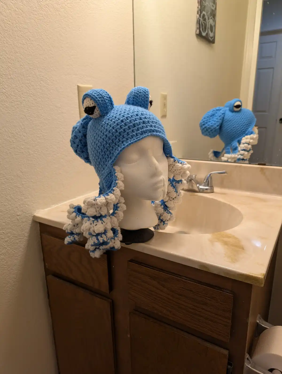 A blue crocheted frog hat with white pom-pom details sits on a white mannequin head placed on a bathroom sink counter. The…
