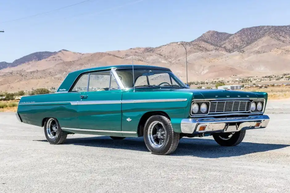 A teal 1960s Ford Fairlane classic car is parked on gravel with mountains in the background. The car has chrome trim, black…