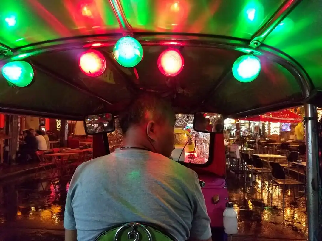 A person viewed from behind sits in a tuk tuk at night, illuminated by alternating red and green lights on the roof. The wet…