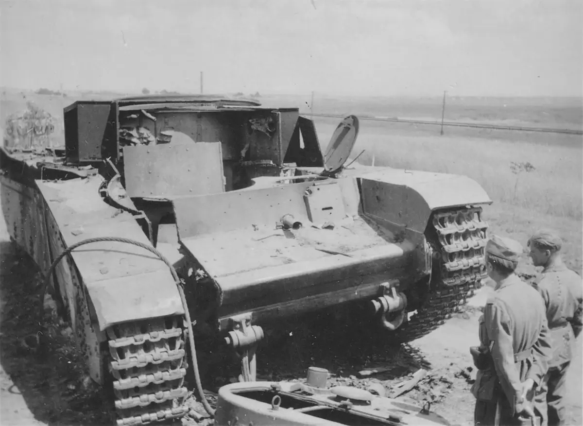 A black-and-white historical photo shows two soldiers standing beside a heavily damaged, open-topped tank with its turret…