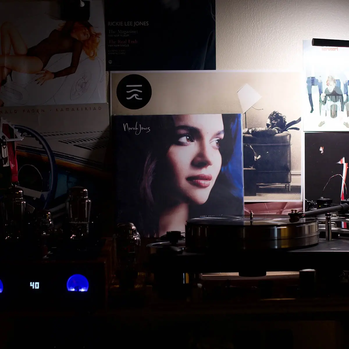 A dimly lit room features a turntable with a vinyl record, surrounded by various album covers on the wall, including Norah…