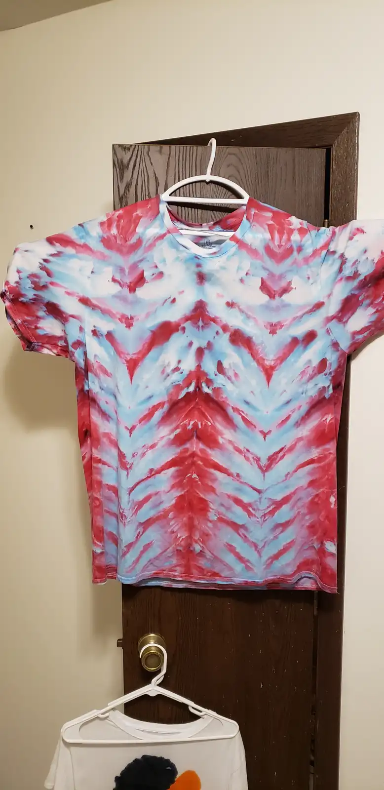 A red, white, and blue tie-dye t-shirt hangs on a white hanger from a dark wooden door, with another white shirt partially…