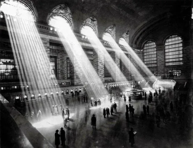 A black-and-white photograph of the grand, vaulted interior of Grand Central Terminal, with dramatic beams of sunlight…