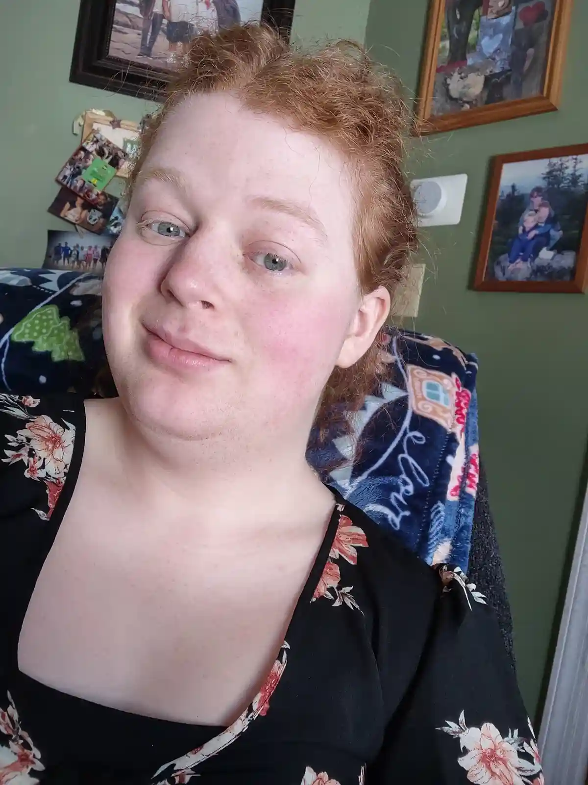 A person with red hair and freckles smiles slightly while sitting in a chair with a patterned blanket draped over their…