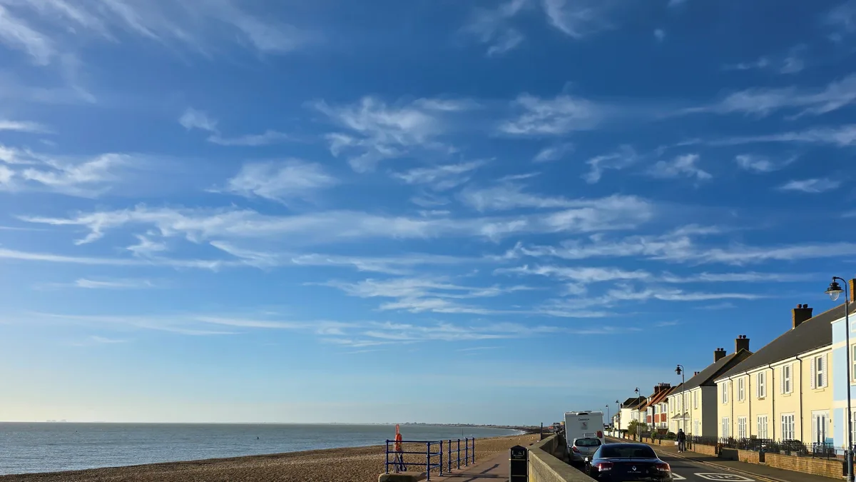 A scenic coastal view shows a bright blue sky with wispy clouds above a calm sea and a pebbled beach. Along the right side…