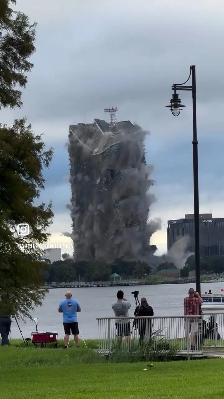 A tall building is collapsing in a controlled demolition, sending a massive cloud of dust and debris skyward. In the…