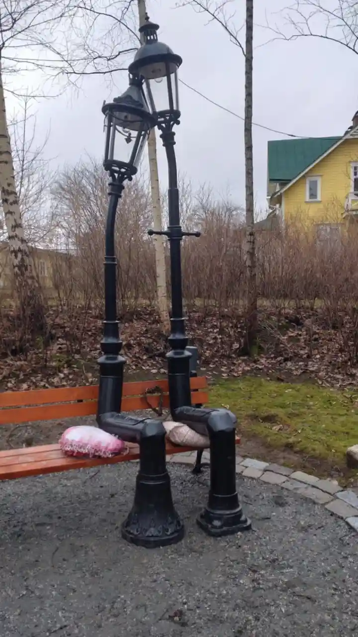Two black ornate street lamps are positioned as if sitting on a wooden park bench, each with a pillow on its 'lap' — one…