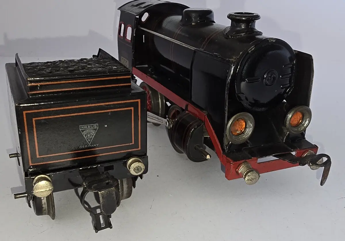 A vintage black toy steam locomotive with red trim and orange headlights is shown pulling a matching black tender car, both…