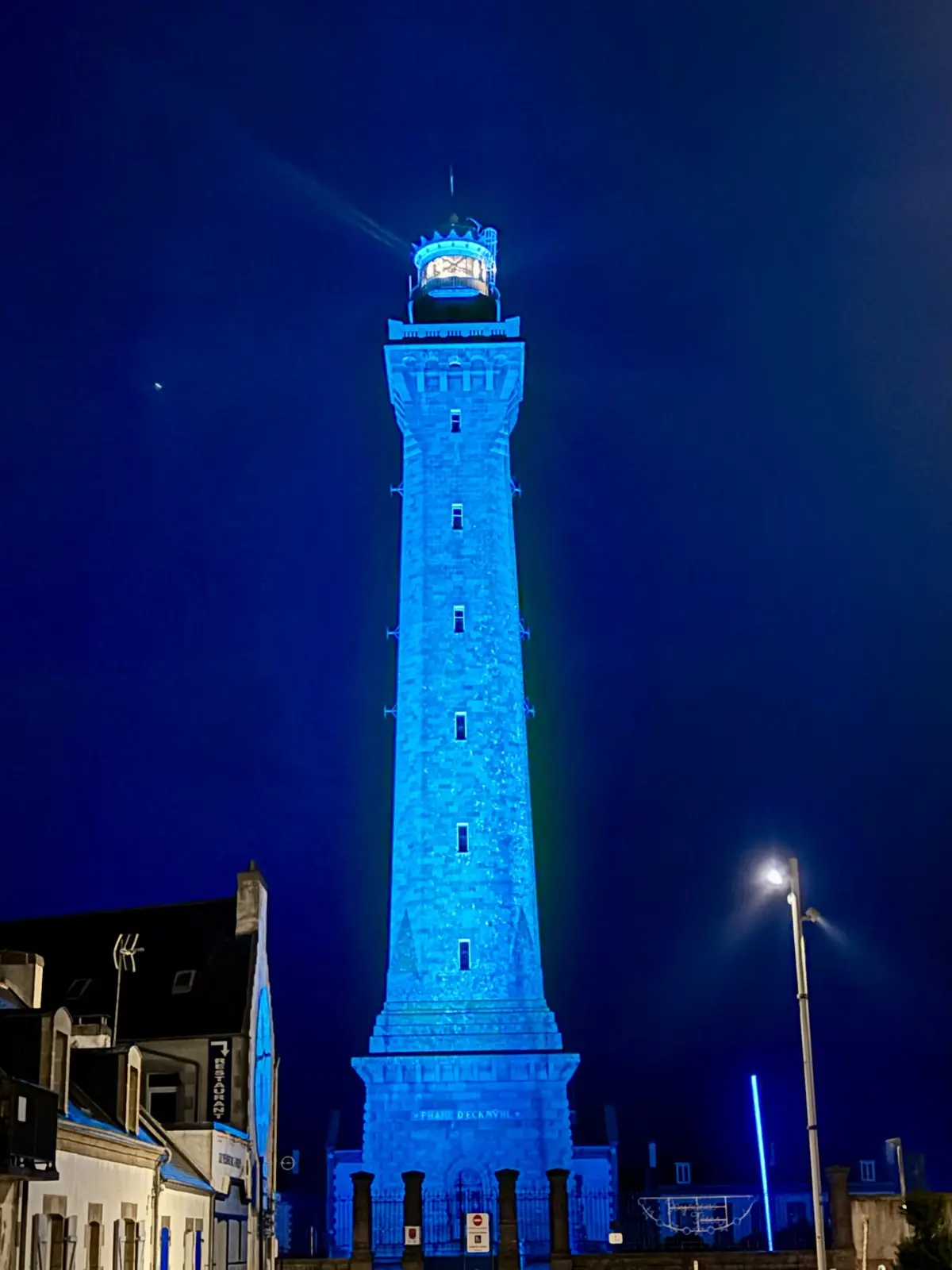 A tall stone lighthouse is illuminated in bright blue light against a dark night sky, with a streetlamp and nearby buildings…