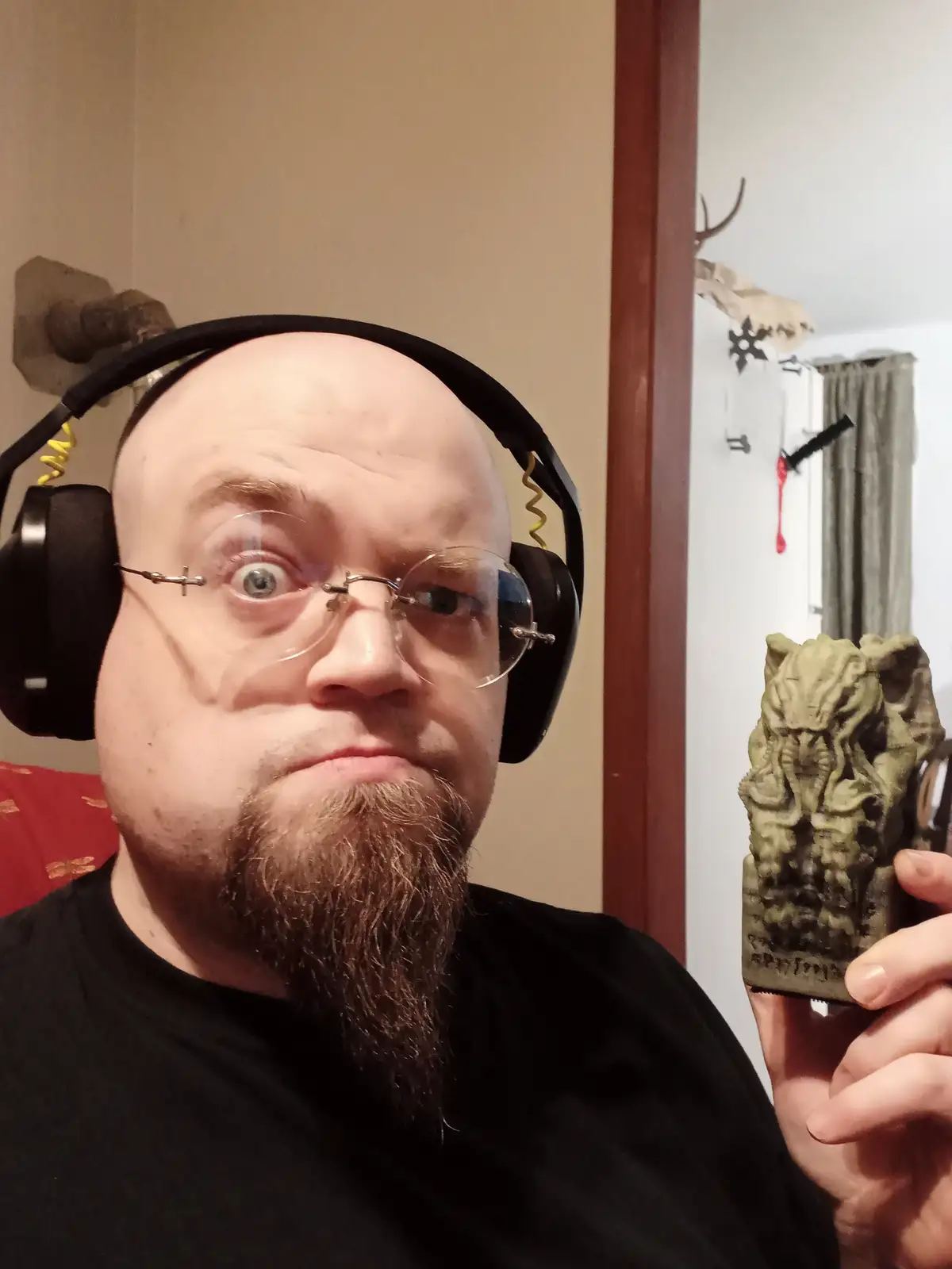A bald man with glasses and a goatee, wearing black headphones, stares with wide eyes at the camera while holding up a…