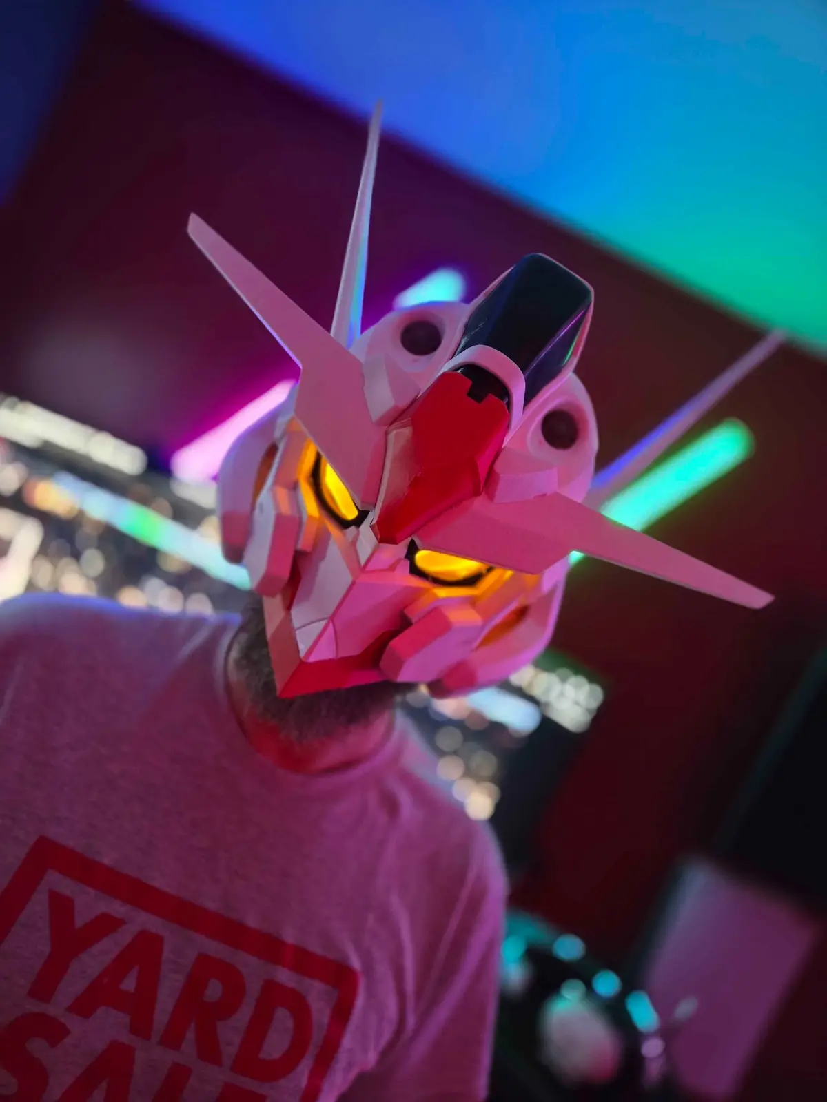 A person wearing a pink and white Gundam-style helmet with glowing yellow eyes, standing in a room with colorful neon…