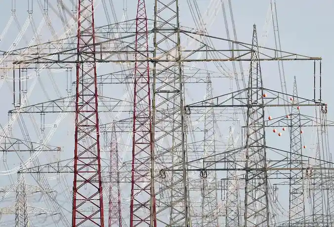A dense, repeating pattern of tall, metal electricity pylons and power lines against a pale sky, with some towers painted…