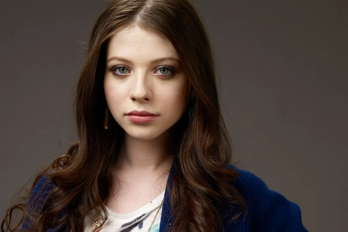 a portrait of a young woman with long brown hair and blue eyes, wearing a blue cardigan over a white shirt, looking directly…