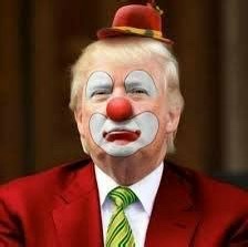 A man with blonde hair wearing a red suit, green tie, and a red clown nose, with white face paint, red lips, and black eye…