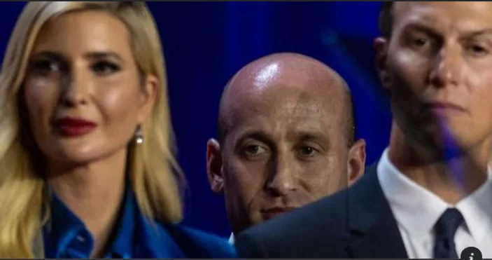 A photo shows three people: a blonde woman in a blue shirt on the left, a bald man in the center, and a man in a suit on the…