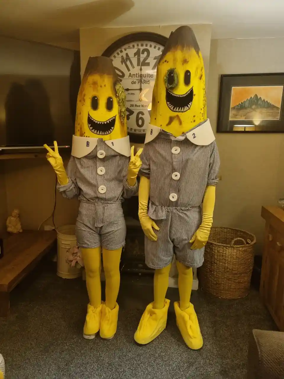 Two people dressed in full banana costumes with cartoonish, toothy faces stand indoors, wearing striped outfits, yellow…