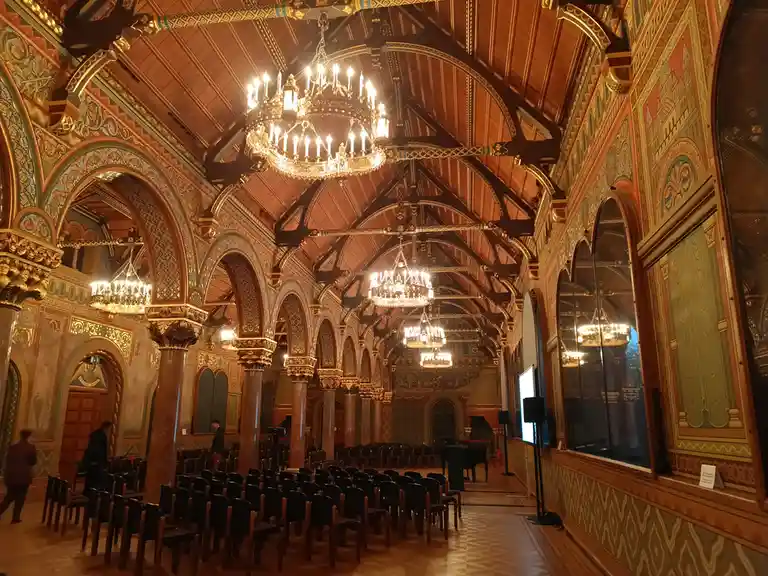 An ornate, grand hall with high arched ceilings, intricate painted murals, and multiple large chandeliers hanging from…