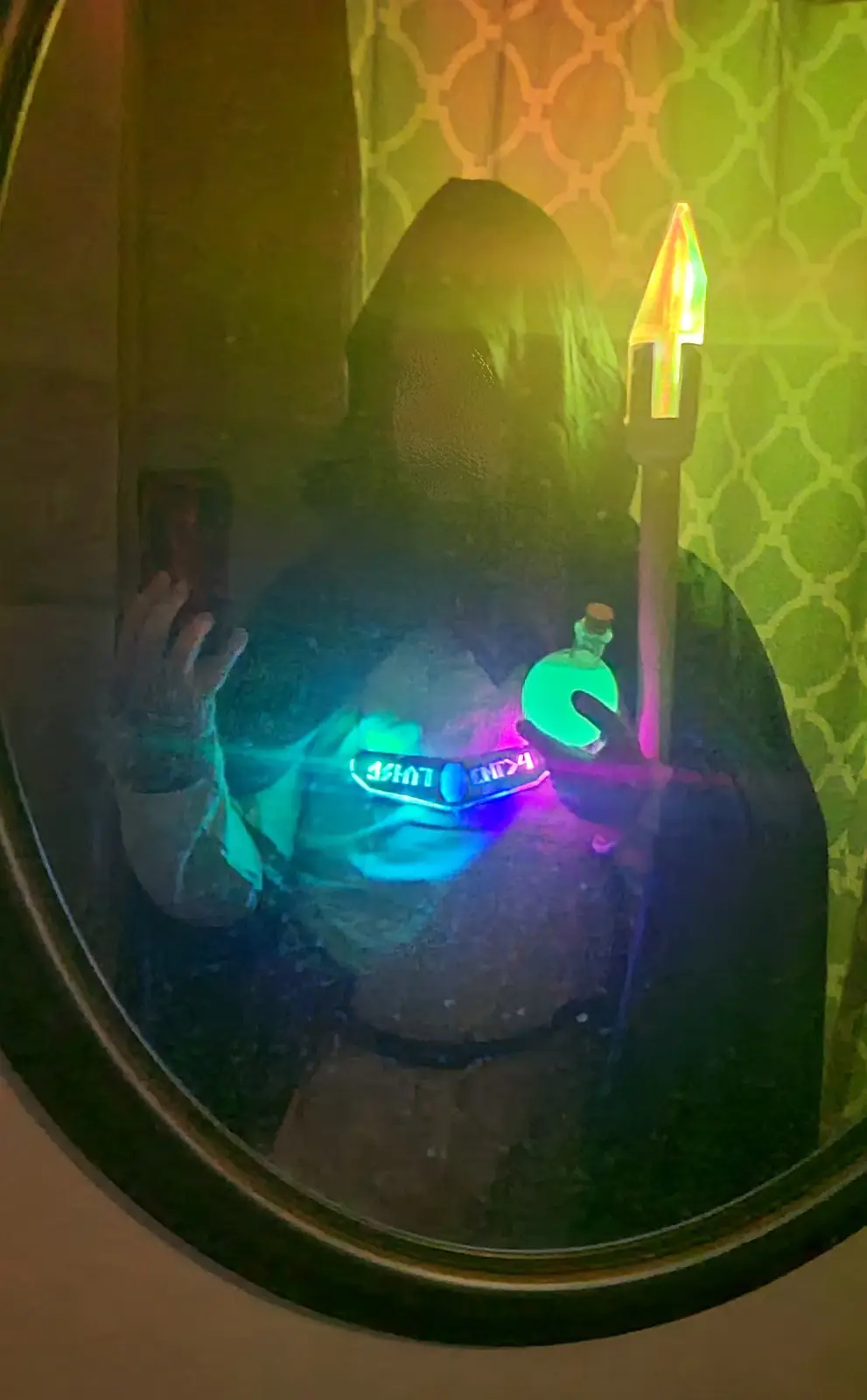 A person in a dark hooded robe takes a mirror selfie while holding a glowing green potion bottle and a glowing staff…
